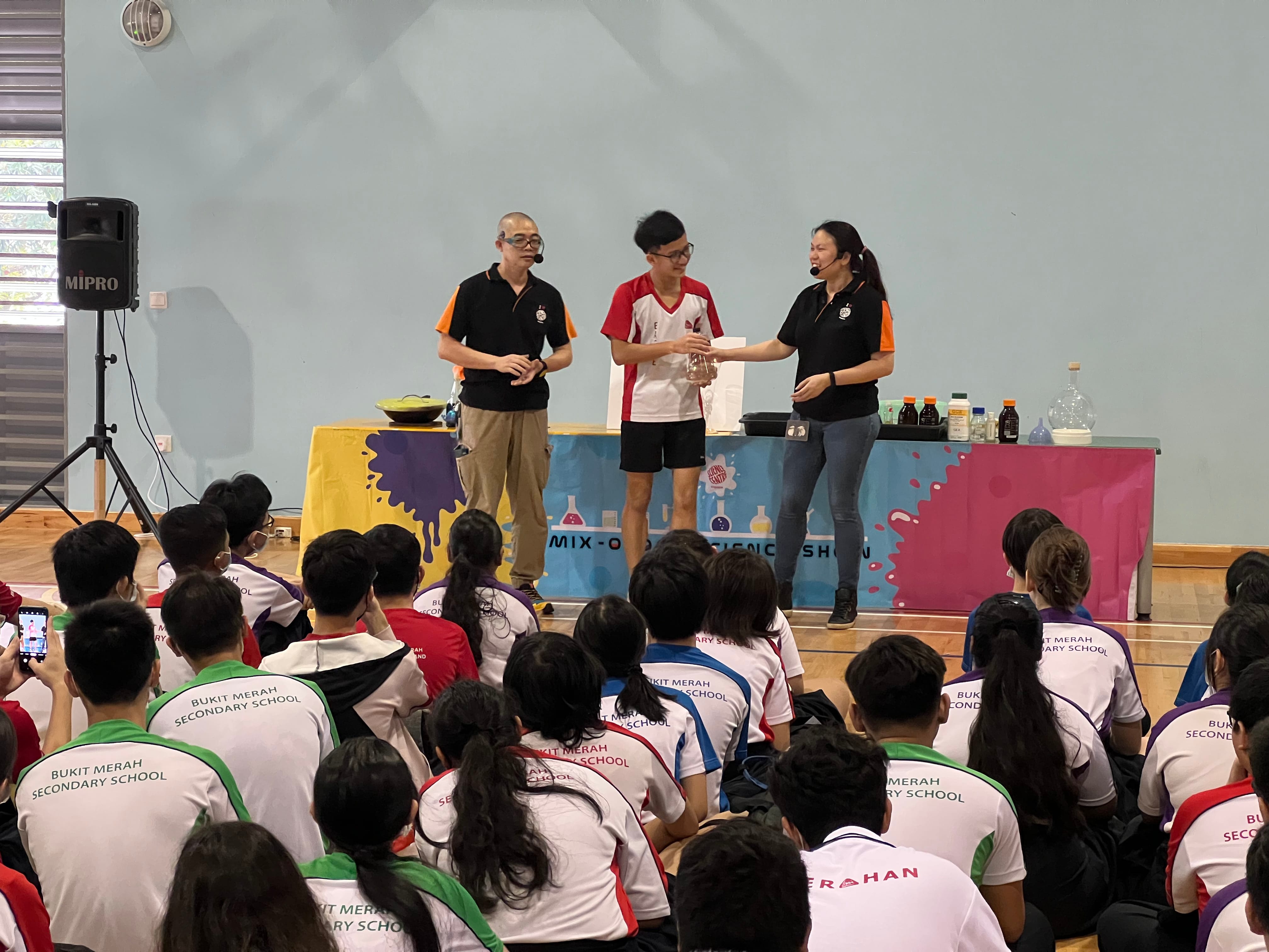 Two presenters and a student perform a science show before a seated crowd of Bukit Merah Secondary School students.